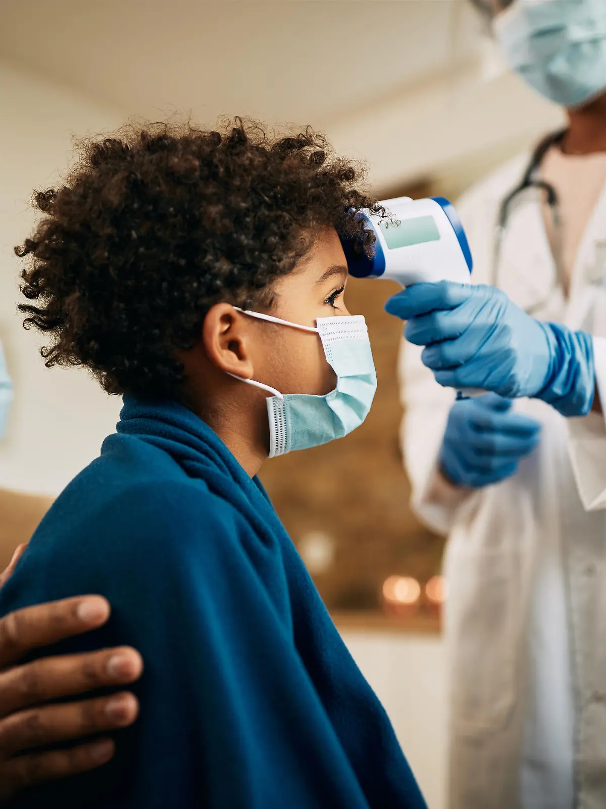 Close-up of pediatrician using infrared thermometer and measuring boy's temperature at home during coronavirus pandemic. Focus is on black boy.