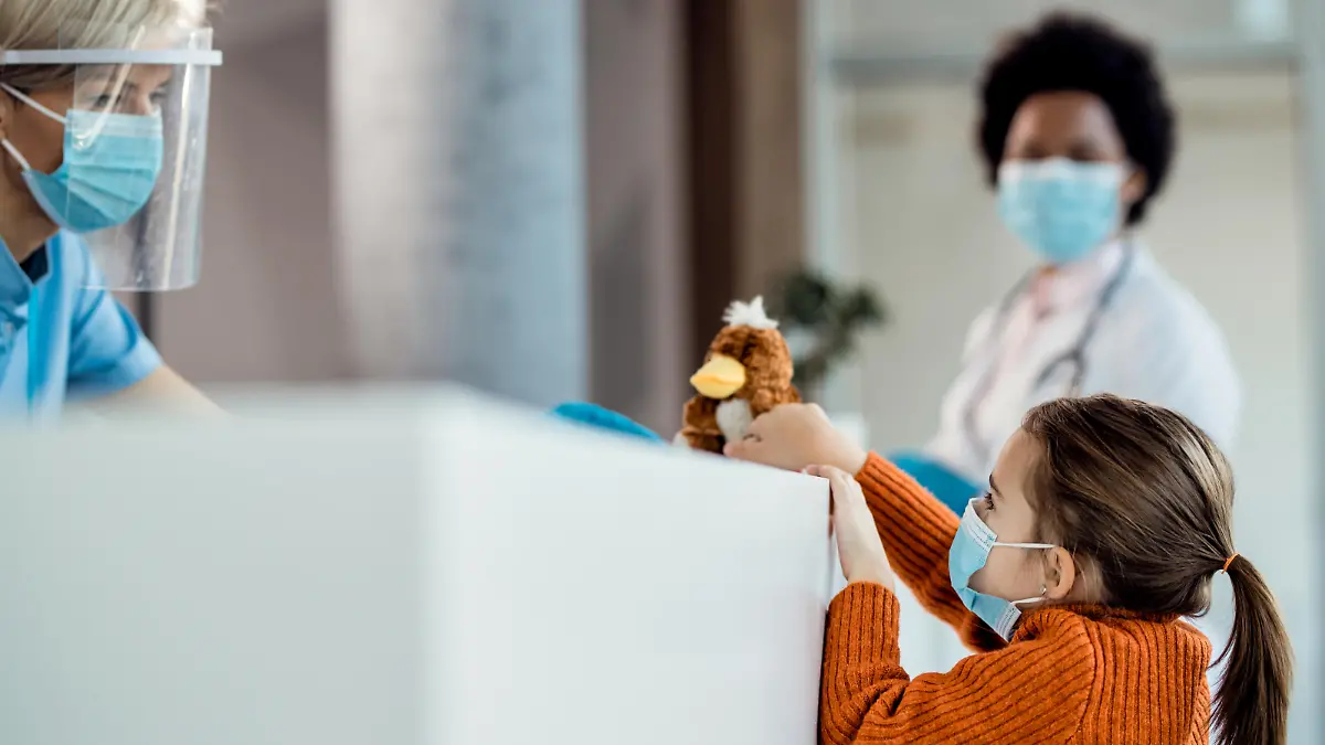 Small girl wearing face mask while communicating with nurse at reception desk at medical clinic.