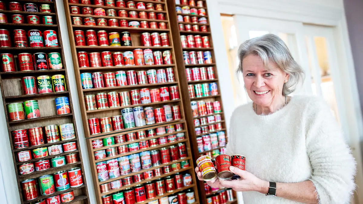 01.02.2022, Niedersachsen, Oldenburg: Gunda Tuchenhagen steht in ihrem Esszimmer vor einer Regalwand mit zahlreichen Tomatenmarkdosen aus verschiedenen Ländern und hält mehrere leere Dosen in den Händen. Die Oldenburgerin hat seit 1983 rund 490 verschiedene Tomatenmarkdosen aus aller Welt gesammelt. (zu dpa "Aus aller Welt: Oldenburgerin sammelt 490 Tomatenmarkdosen") Foto: Hauke-Christian Dittrich/dpa - ACHTUNG: Nur zur redaktionellen Verwendung im Zusammenhang mit der aktuellen Berichterstattung über diesen Bericht. +++ dpa-Bildfunk +++