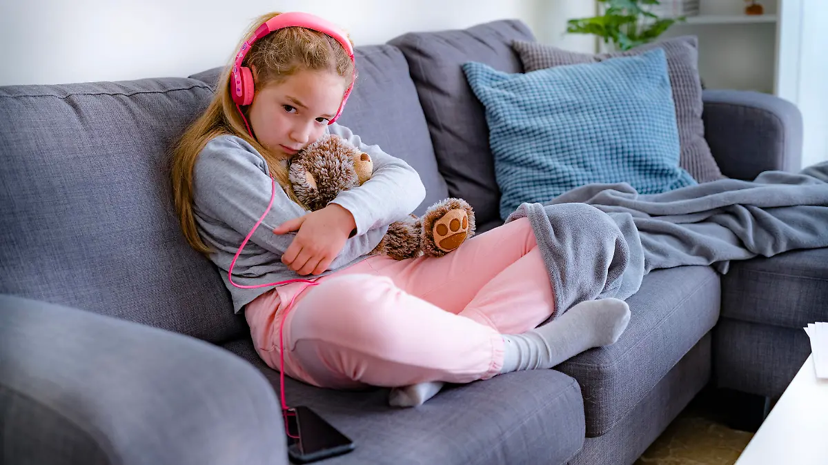 Little girl in home clothes sitting on sofa with her Teddy Bear, listening music, bored at home during quarantine due Coronavirus pandemic. Her face express sadness, loneliness, confusion, emotional stress, worry and other negative emotions. High resolution 42Mp studio digital capture taken with SONY A7rII and Zeiss Batis 40mm F2.0 CF lens