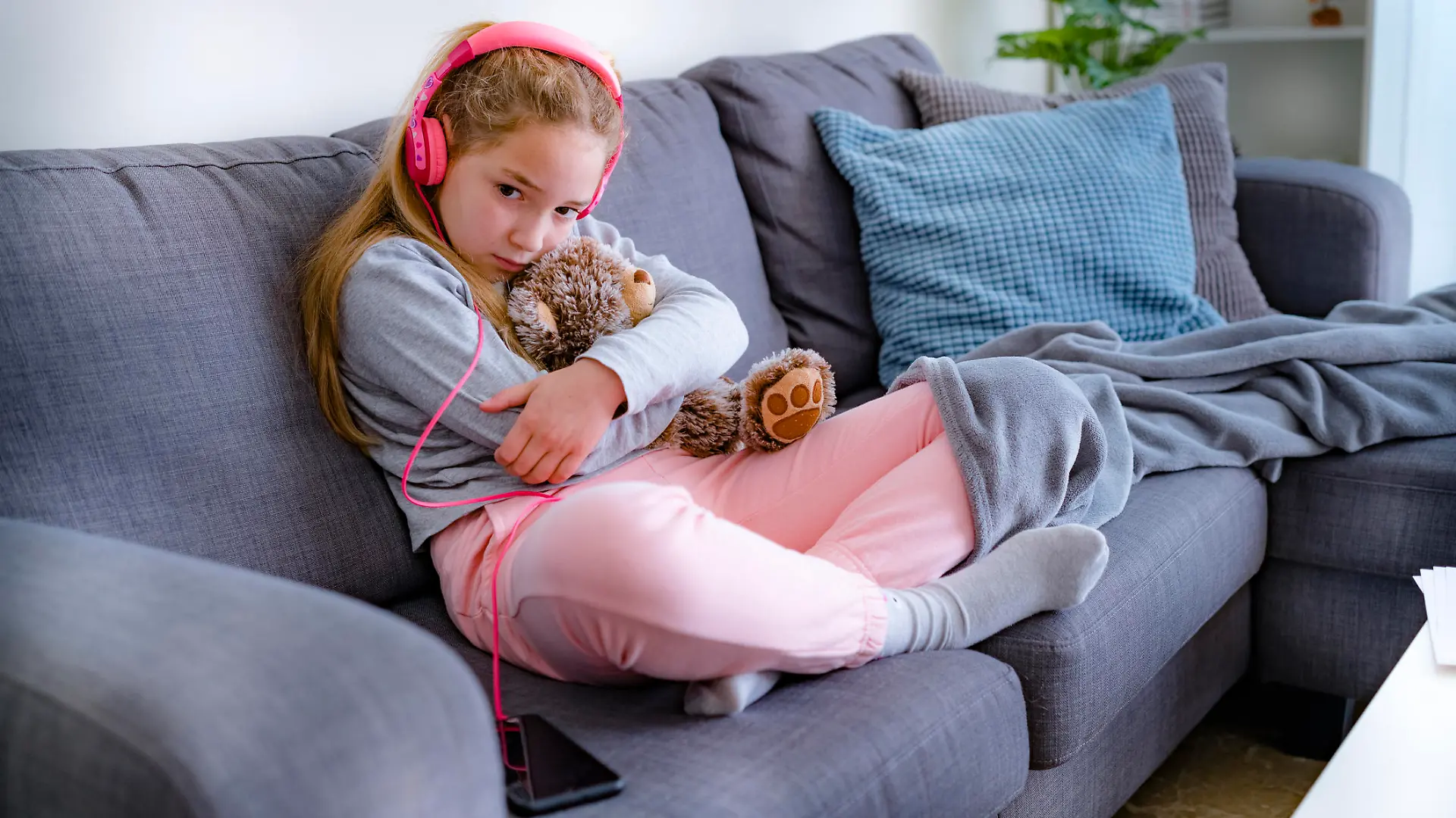 Little girl in home clothes sitting on sofa with her Teddy Bear, listening music, bored at home during quarantine due Coronavirus pandemic. Her face express sadness, loneliness, confusion, emotional stress, worry and other negative emotions. High resolution 42Mp studio digital capture taken with SONY A7rII and Zeiss Batis 40mm F2.0 CF lens