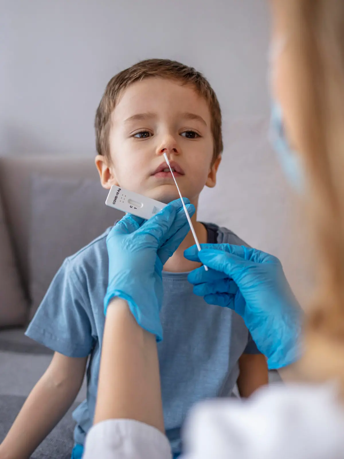 Boy sitting on sofa while going through PCR testing at home due to COVID-19 pandemic. Female doctor using cotton swab while PCR testing small boy at rapid COVID-19 test at home.