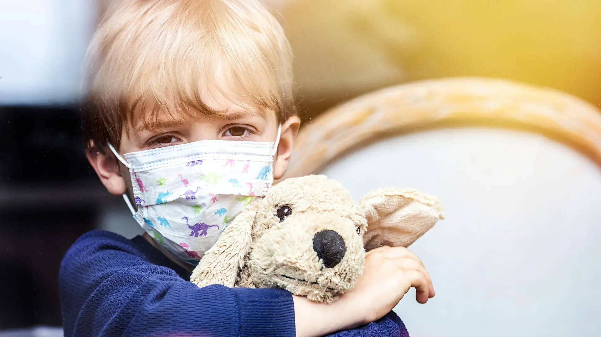 Caucasian sad Little child boy looking through a window using a surgical mask holding a toy dog
