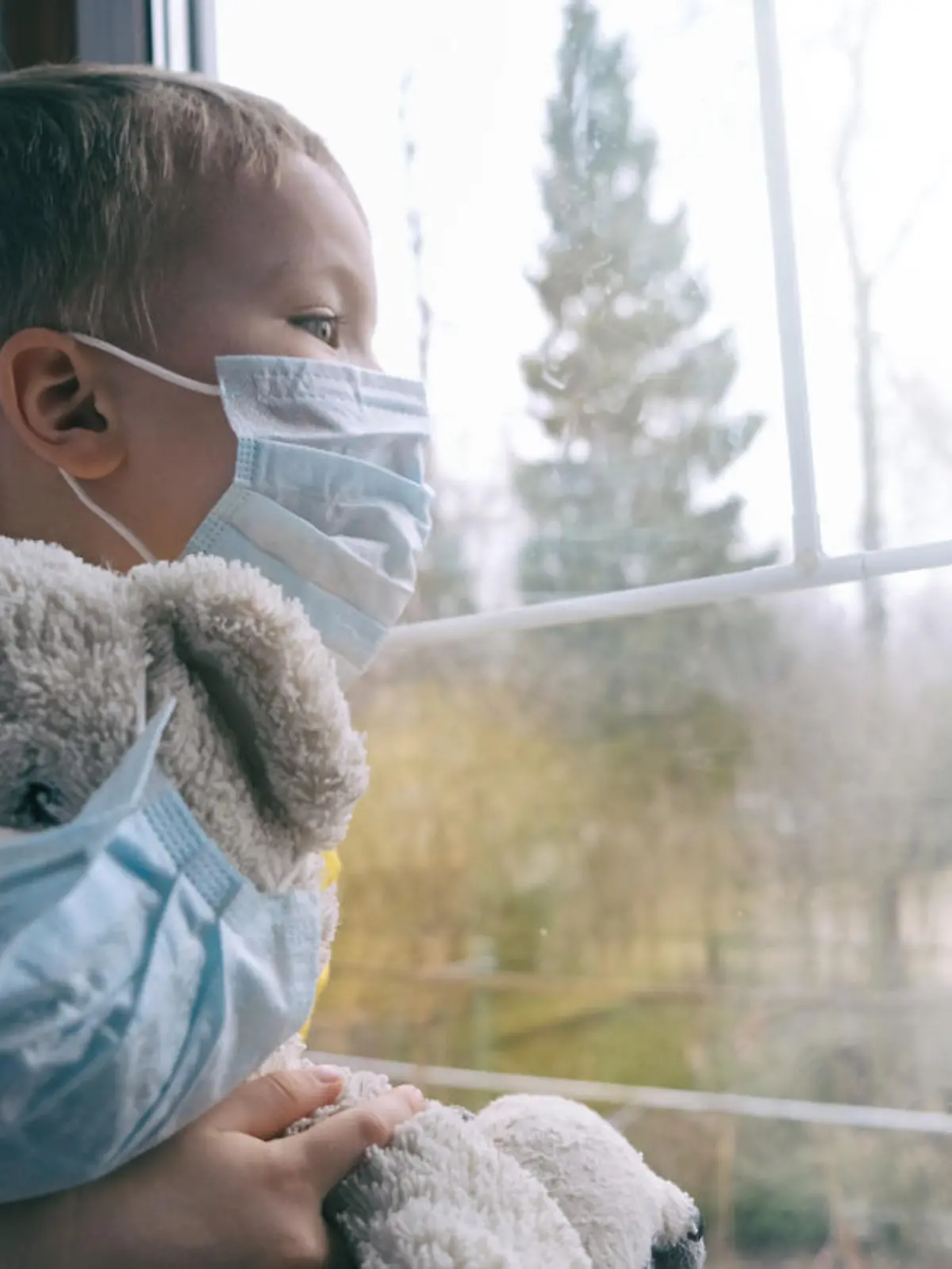 Sad illness child on home quarantine. Boy and his teddy bear both in protective medical masks sits on windowsill and looks out window. Virus protection, coronavirus pandemic, prevention epidemic.
