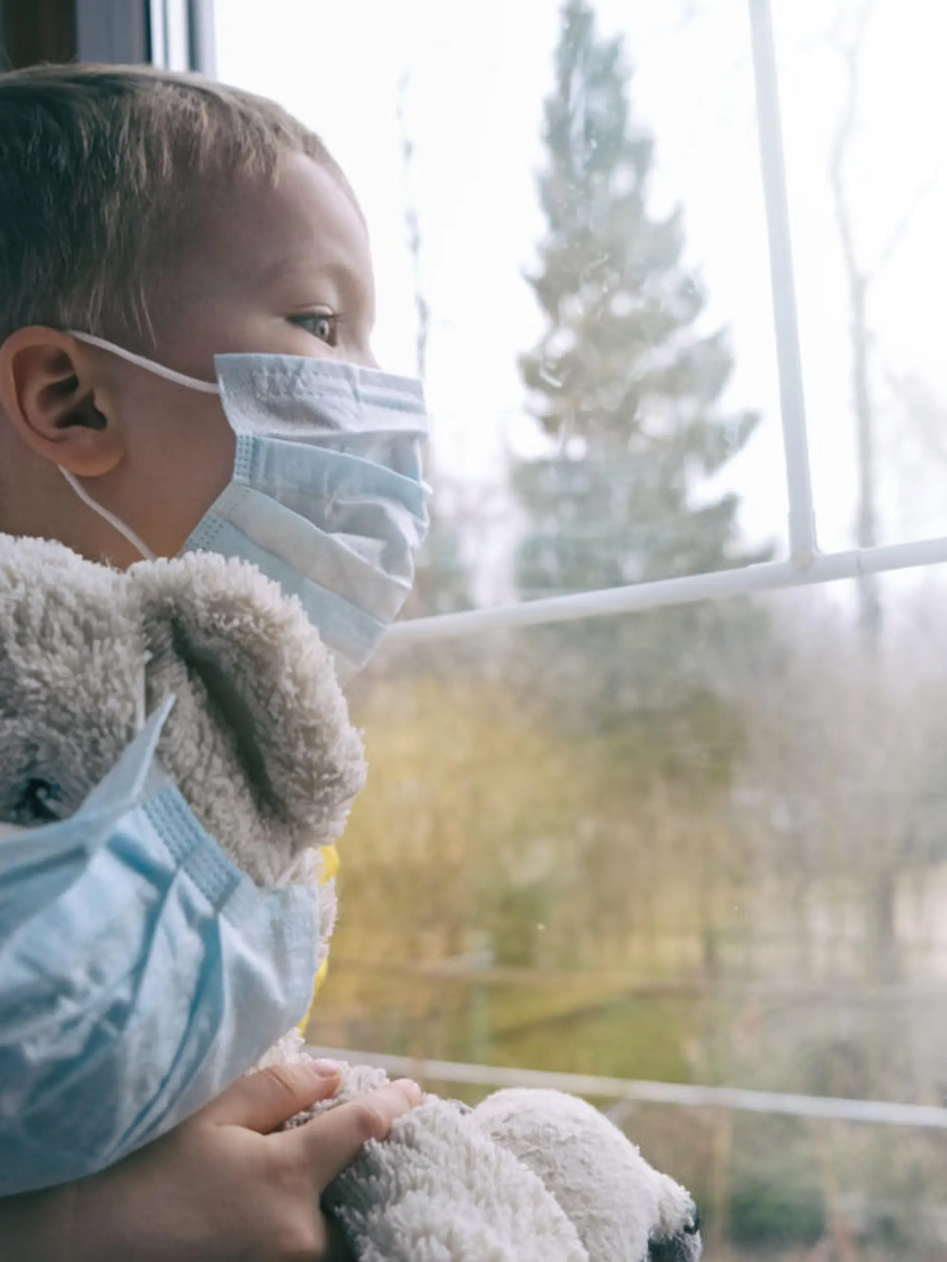 Sad illness child on home quarantine. Boy and his teddy bear both in protective medical masks sits on windowsill and looks out window. Virus protection, coronavirus pandemic, prevention epidemic.