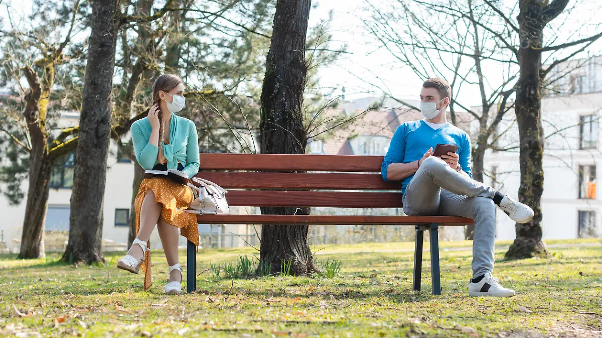 Woman and man with face mask in social distancing flirting sitting on a park bench