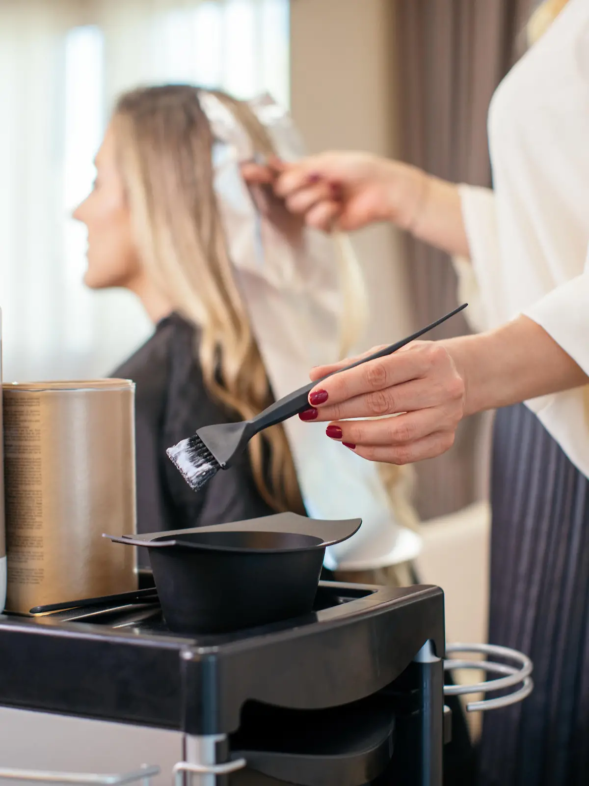 Blonde woman dyeing her hair at the salon. She is sitting and the hairdresser/hairstylist is putting hair color on her hair. This is regular hair care treatment in customers favorite salon.