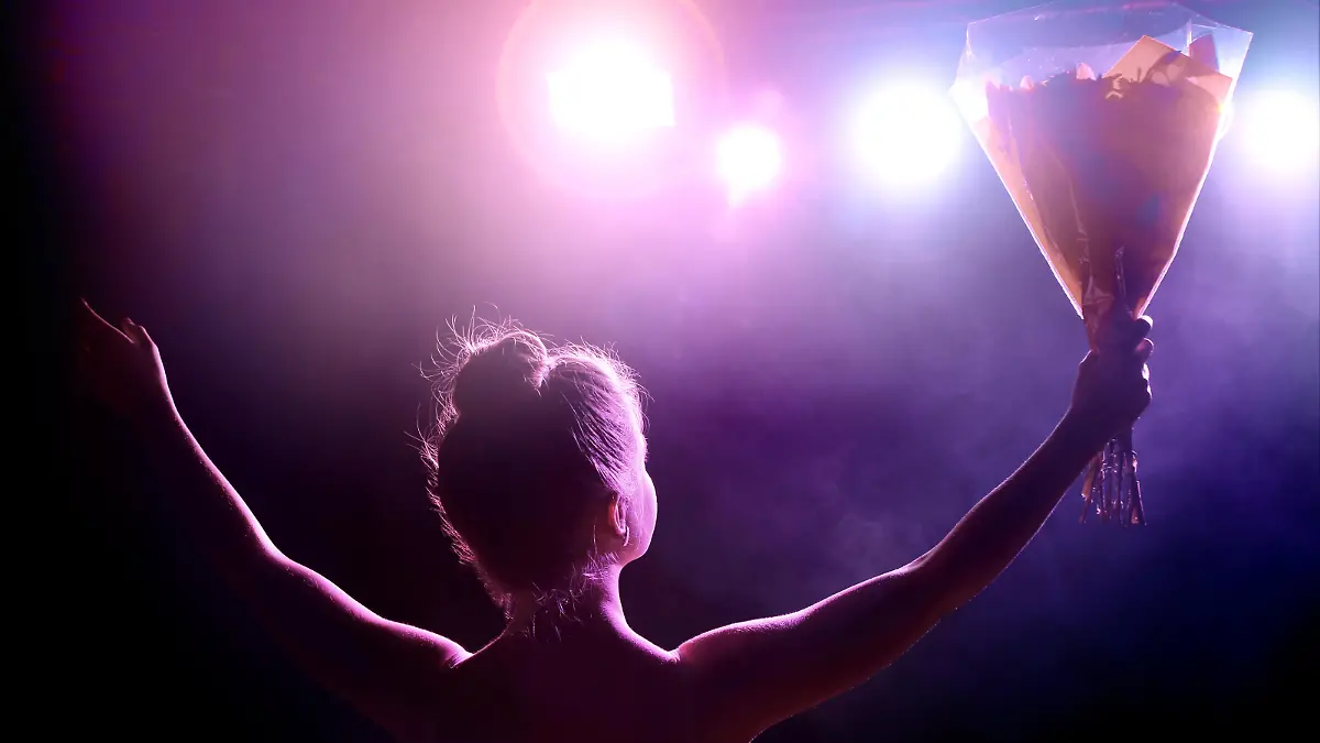 Horizontal composition color photography of young woman artist (singer or classical dancer) receives a bouquet of flowers on stage at the end of the spectacle as a thank you, and saluting the public. Rear view from backstage with spotlight in camera creating lens flare.