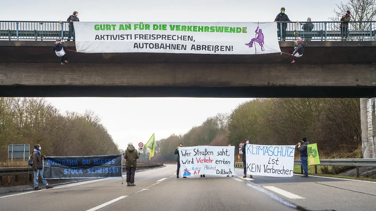 23.01.2022, Hessen, Künzell: Aktivisten mit Transparenten stehen auf der gesperrten Fahrbahn der Autobahn A7 am Rande von Künzell bei Fulda, während oben an einer Bücke eine genehmigte Abseilaktion stattfindet. Mit der Aktion soll für eine echte Verkehrswende, den sofortigen Stopp des Straßenbaus, einen konsequenten Klimaschutz und gegen Strafen für Autobahnaktionen geworben werden. Foto: Frank Rumpenhorst/dpa/Frank Rumpenhorst/dpa +++ dpa-Bildfunk +++