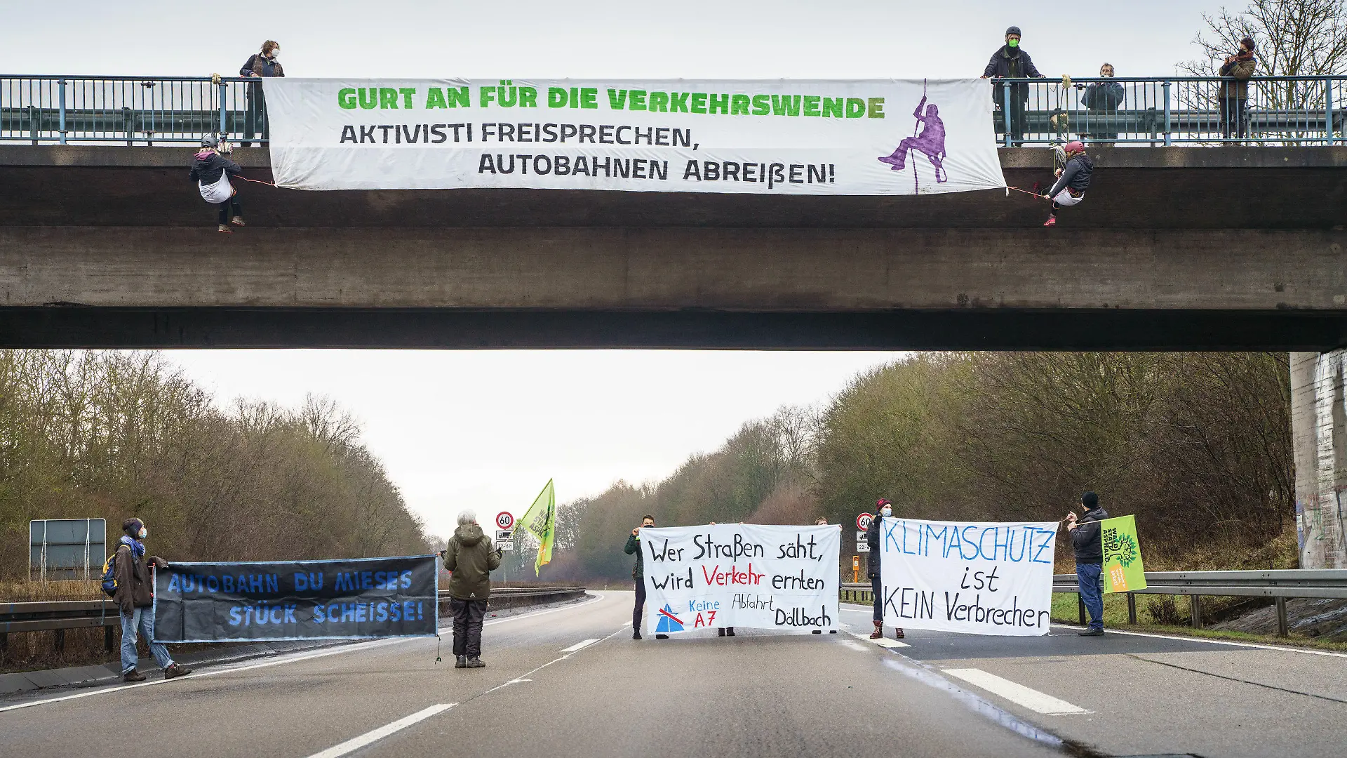 23.01.2022, Hessen, Künzell: Aktivisten mit Transparenten stehen auf der gesperrten Fahrbahn der Autobahn A7 am Rande von Künzell bei Fulda, während oben an einer Bücke eine genehmigte Abseilaktion stattfindet. Mit der Aktion soll für eine echte Verkehrswende, den sofortigen Stopp des Straßenbaus, einen konsequenten Klimaschutz und gegen Strafen für Autobahnaktionen geworben werden. Foto: Frank Rumpenhorst/dpa/Frank Rumpenhorst/dpa +++ dpa-Bildfunk +++