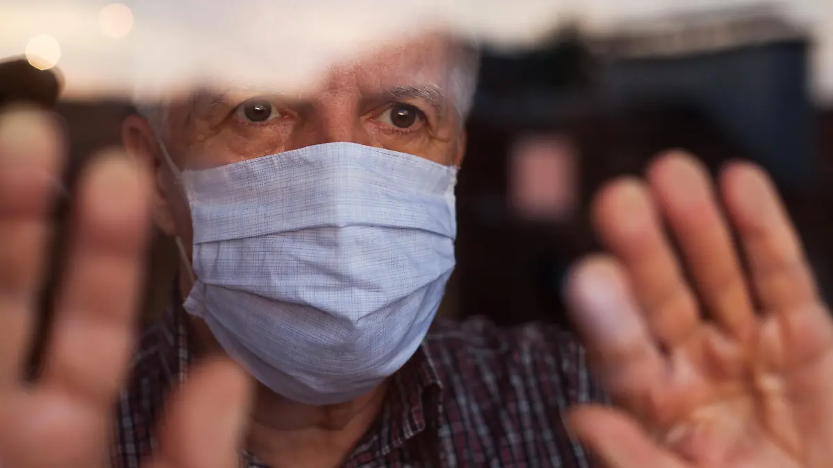 Elderly caucasian man wearing hand made protective face mask, in nursing care home, looking outside window, sadness, stress & hope in his eyes, self isolation due to global COVID-19 Coronavirus pandemic