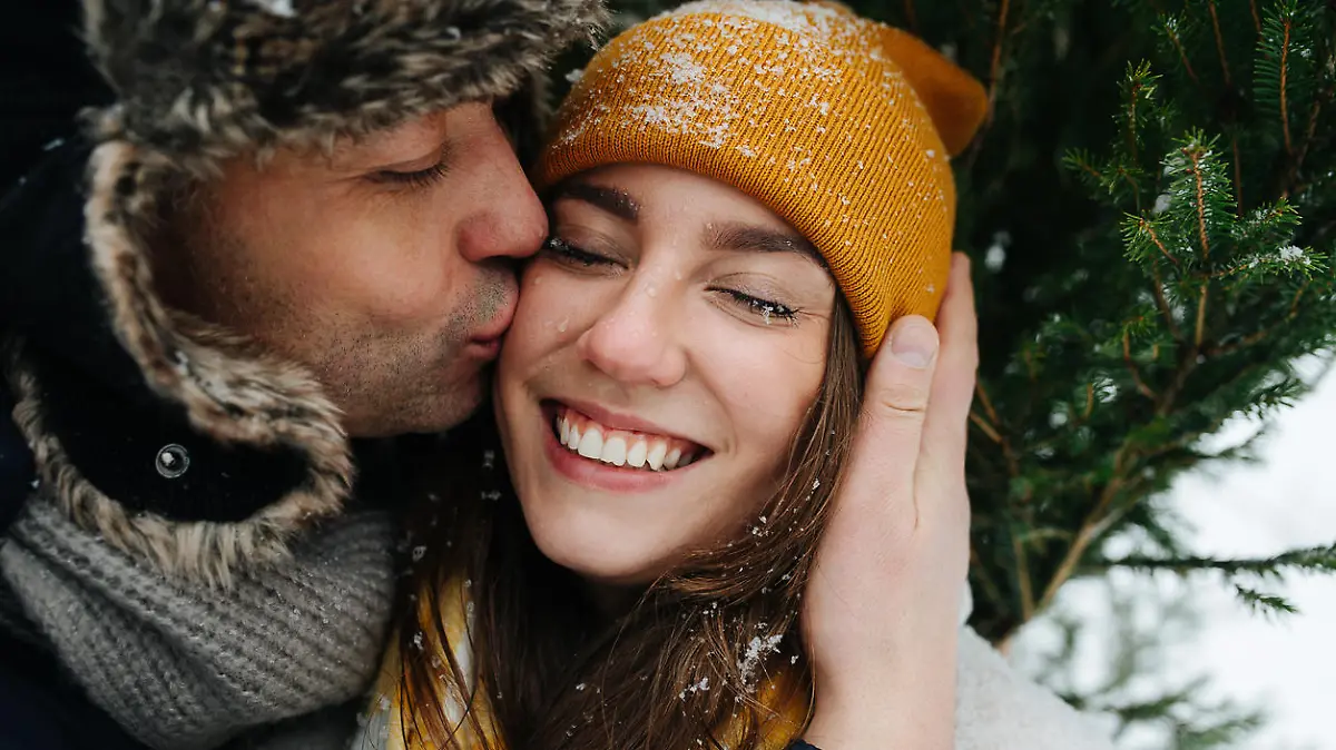 Happy man kissing his smiling woman in a cheek in front of a fir tree in the winter. Close up.