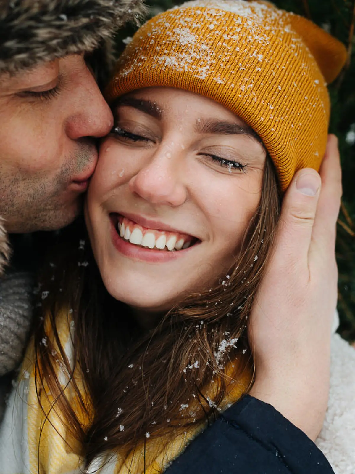 Happy man kissing his smiling woman in a cheek in front of a fir tree in the winter. Close up.