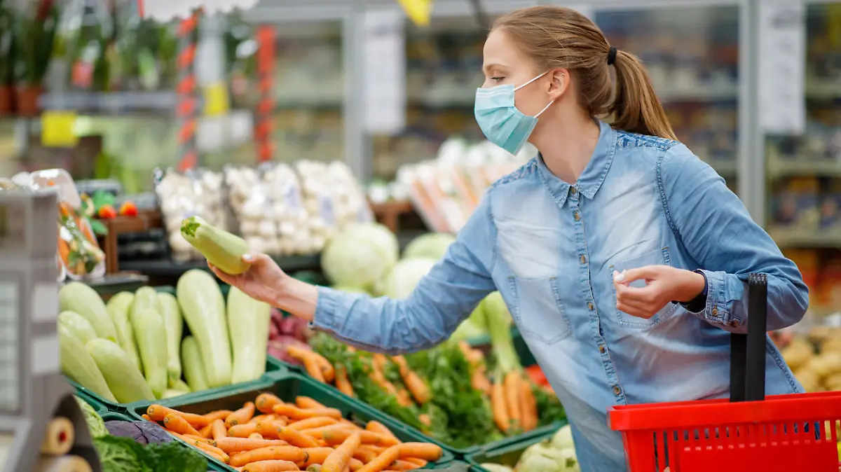 Young woman choosing fresh zucchini from produce section in supermarket, standing next to produce section, she is wearing disposable face mask amid Covid-19 pandemia