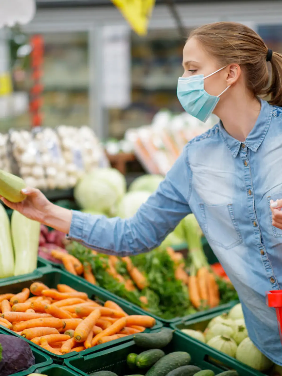 Young woman choosing fresh zucchini from produce section in supermarket, standing next to produce section, she is wearing disposable face mask amid Covid-19 pandemia