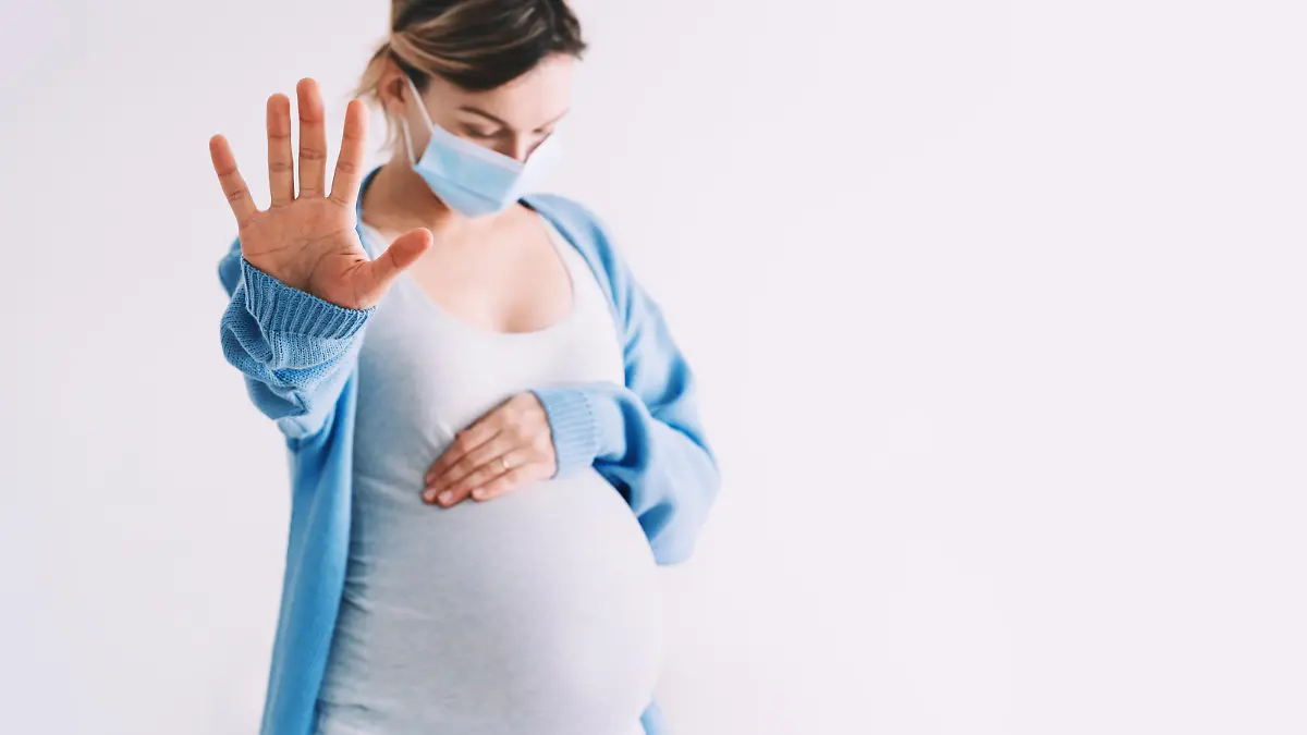 Pregnancy and childbirth during Infections. Pregnant woman in medical mask hugging belly showing STOP gesture. Protection and prevention during viruses, mother care of her and unborn baby health.