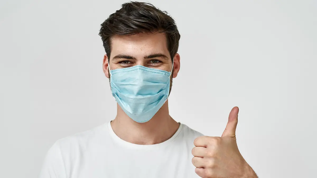 Covid-19 protection. Young man wearing medical mask showing the thumbs up while standing against grey background. Coronavirus prevention concept