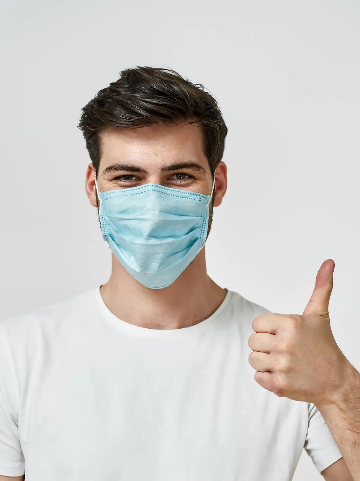 Covid-19 protection. Young man wearing medical mask showing the thumbs up while standing against grey background. Coronavirus prevention concept