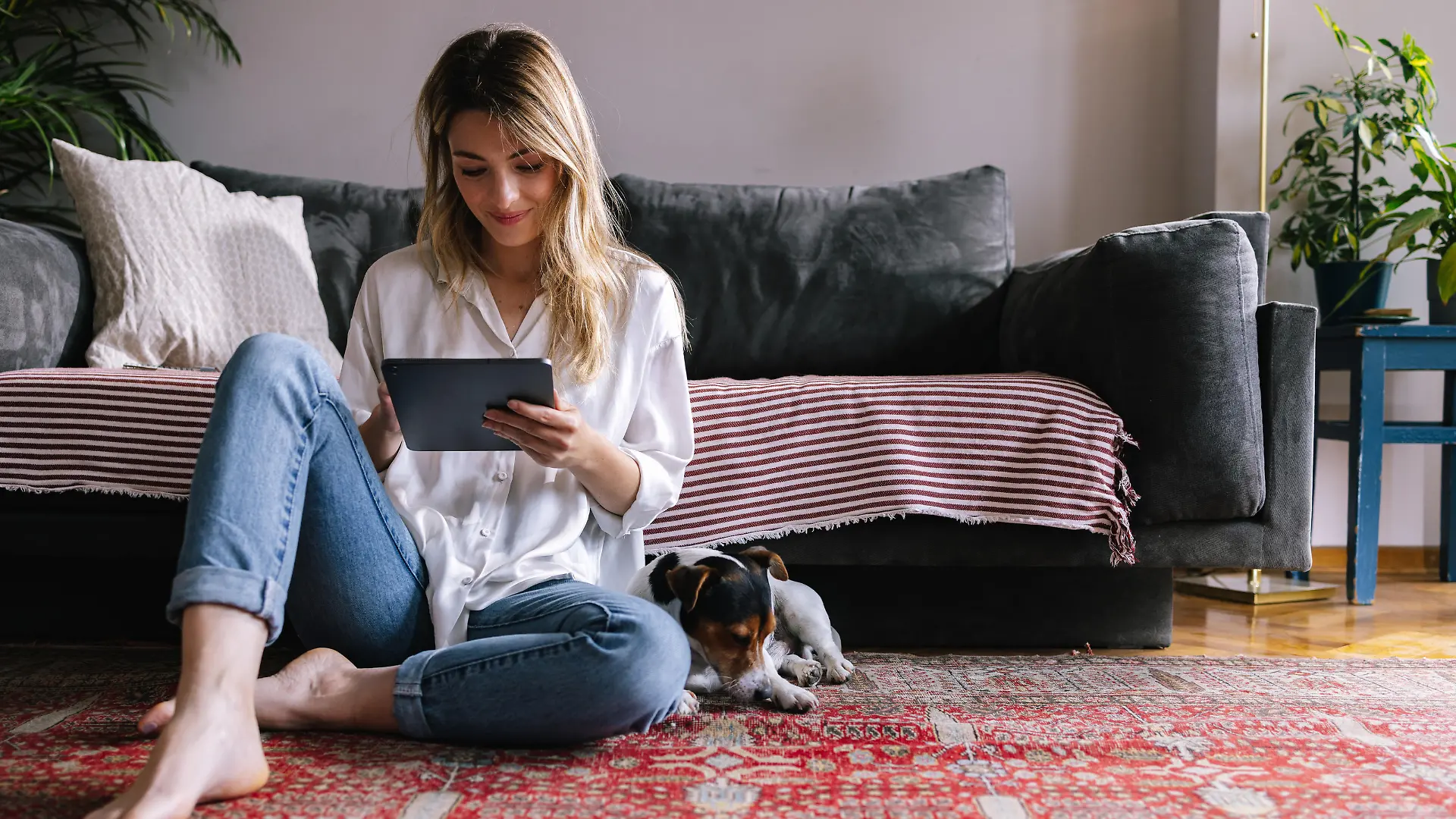 Attractive blonde Caucasian sitting on the floor of her living room, enjoying her free time by reading a book on a Kindle device. Her dog is lying next to her.