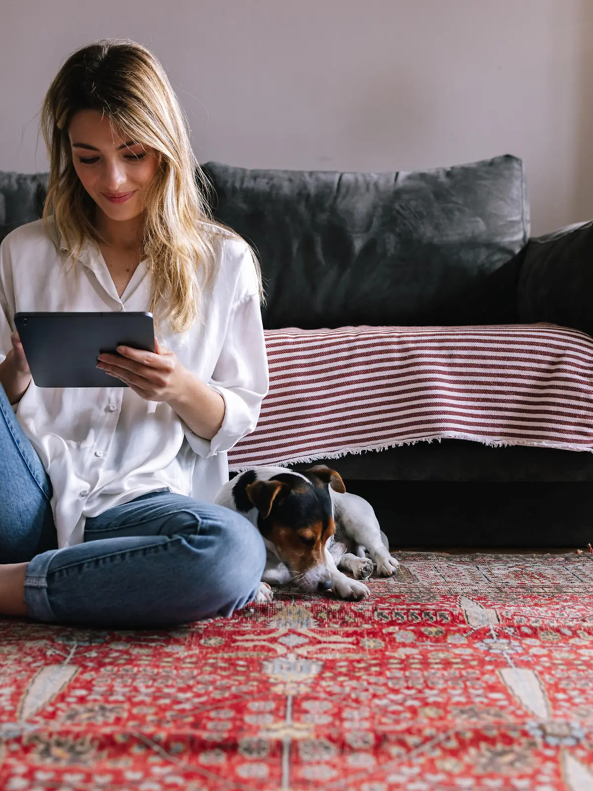 Attractive blonde Caucasian sitting on the floor of her living room, enjoying her free time by reading a book on a Kindle device. Her dog is lying next to her.