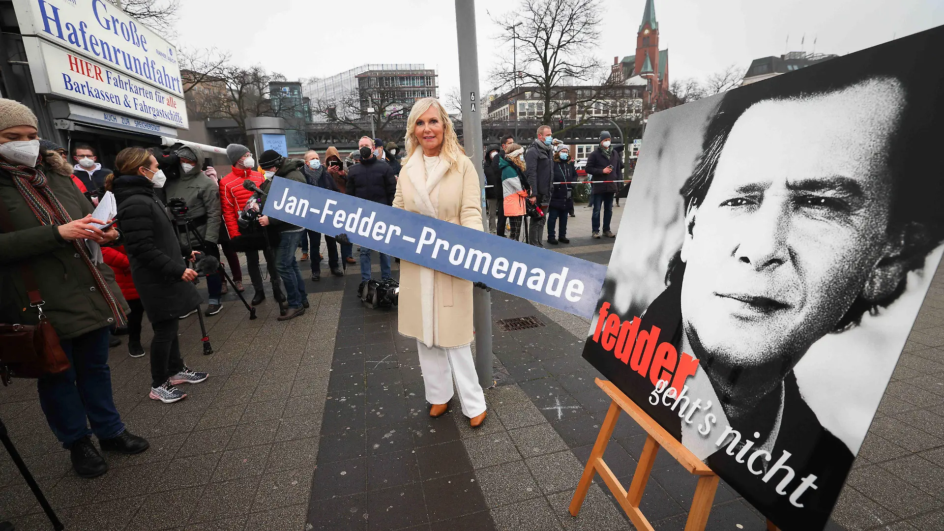 14.01.2022, Hamburg: Marion Fedder (M), Witwe von Jan Fedder, hält ein Schild mit dem Text «Jan-Fedder-Promenade» bei der Einweihung der Jan-Fedder-Promenade an den Landungsbrücken. Die Elbpromenade zwischen Landungsbrücken und Baumwall wurde am Freitag nach dem 2019 gestorbenen Hamburger Volksschauspieler benannt. Foto: Christian Charisius/dpa +++ dpa-Bildfunk +++