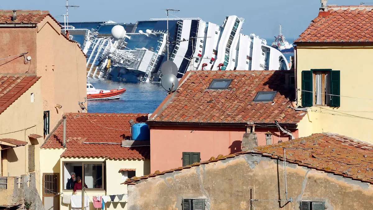 FILE— A woman hangs her laundry as the grounded cruise ship Costa Concordia is seen in the background, off the Tuscan island of Isola del Giglio, Italy, Saturday, Jan. 21, 2012. Italy on Thursday, Jan. 13, 2022, is marking the 10th anniversary of the Concordia disaster with a daylong commemoration, honoring the 32 people who died but also the extraordinary response by the residents of Giglio who took in the 4,200 passengers and crew from the ship on that rainy Friday night and then lived with the Concordia carcass for another two years before it was hauled away for scrap.(AP Photo/Pier Paolo Cito)