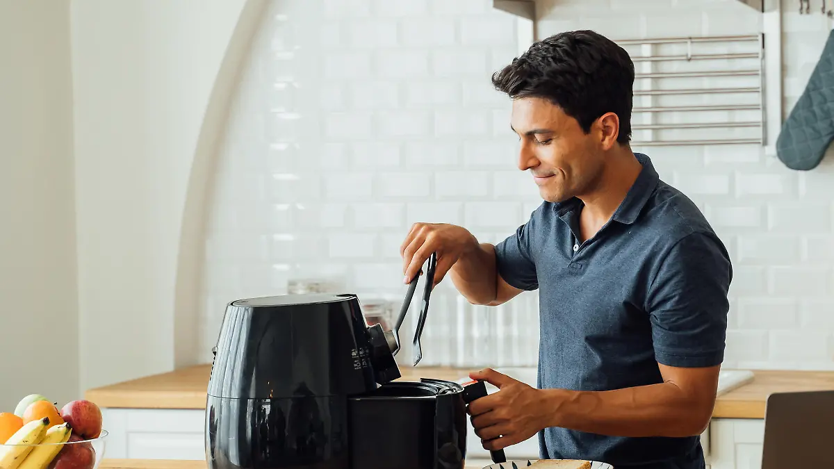 Handsome young caucasian man looking at instructions online on laptop on how to use modern air fryer to cook healthy food without using oil at home in kitchen