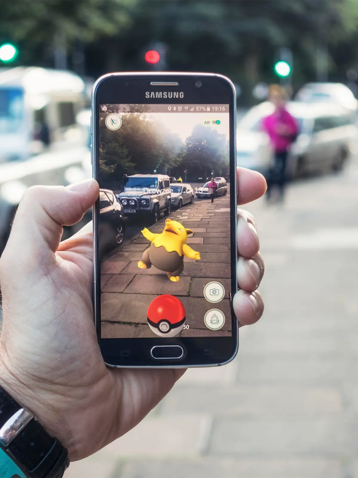 Edinburgh, UK - July 18, 2016: Closeup of a man holding a Samsung S6 smartphone, playing Pokemon Go with the game's augmented reality superimposing a character onto the pavement surface, as a person approaches in the distance.
