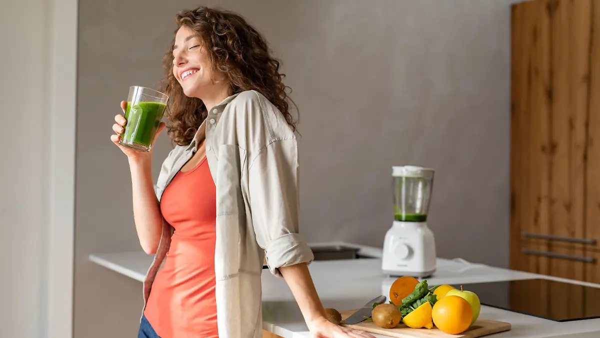 Happy young woman standing in kitchen and holding glass of detox juice. Cheerful girl drinking healthy smoothie at home. Beautiful smiling woman drinking green vegetable smoothie.