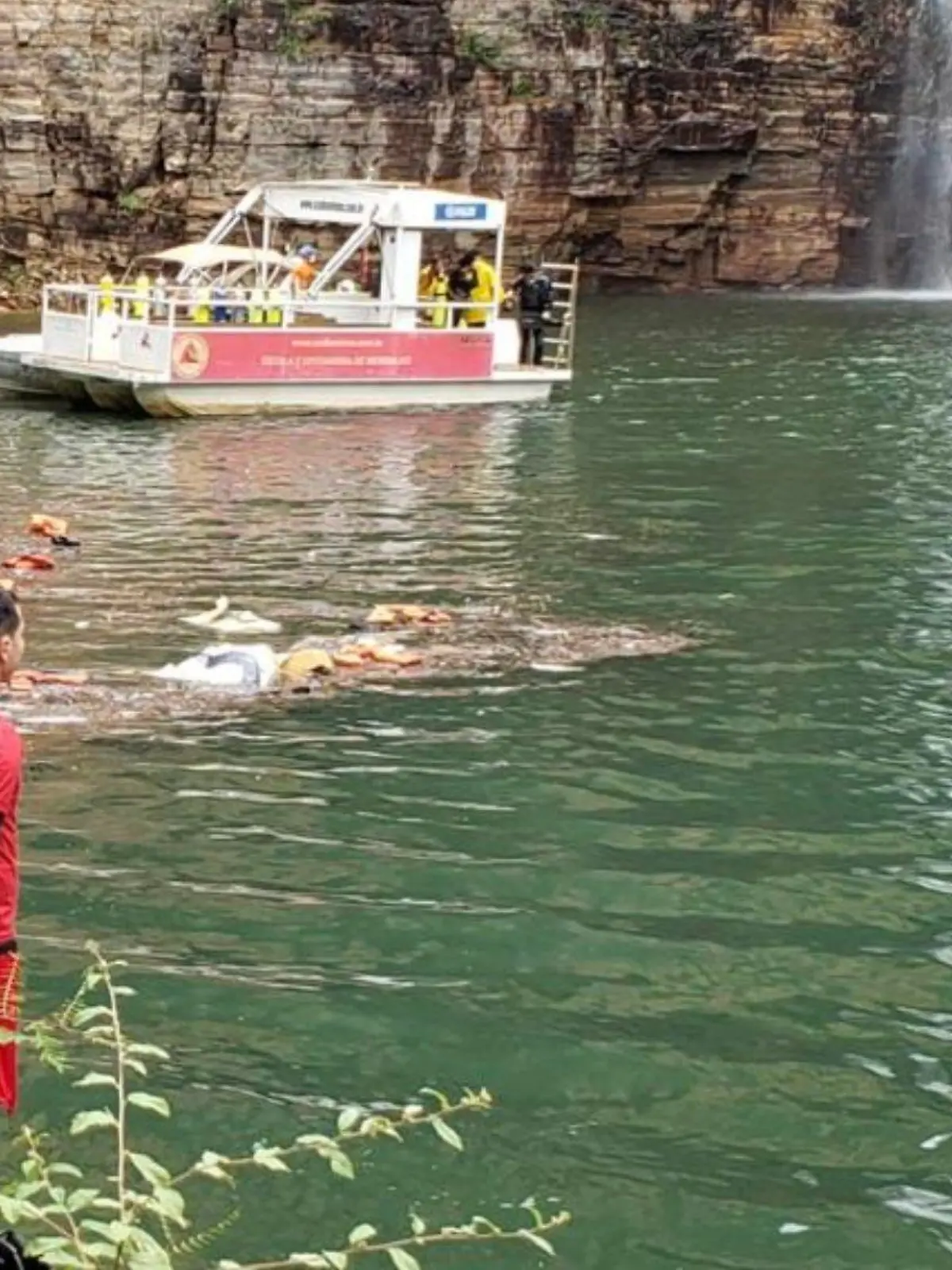 Mann steht mit Rettungsweste vor Felswand, Taucher suchen im See nach Leichen