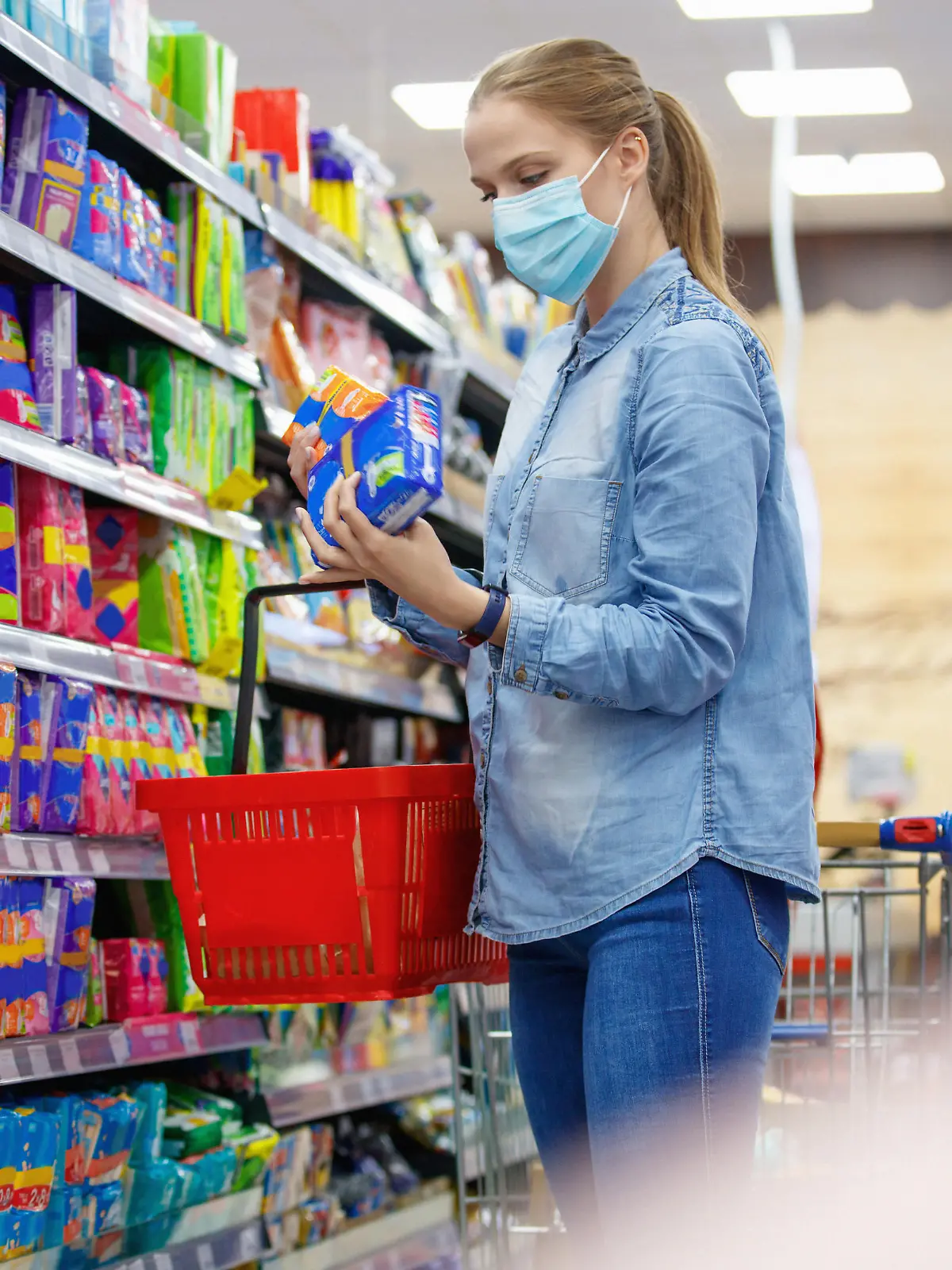 Young woman wearing protective face mask, choosing sanitary pads in supermarket aisle, COVID-19 pandemic era