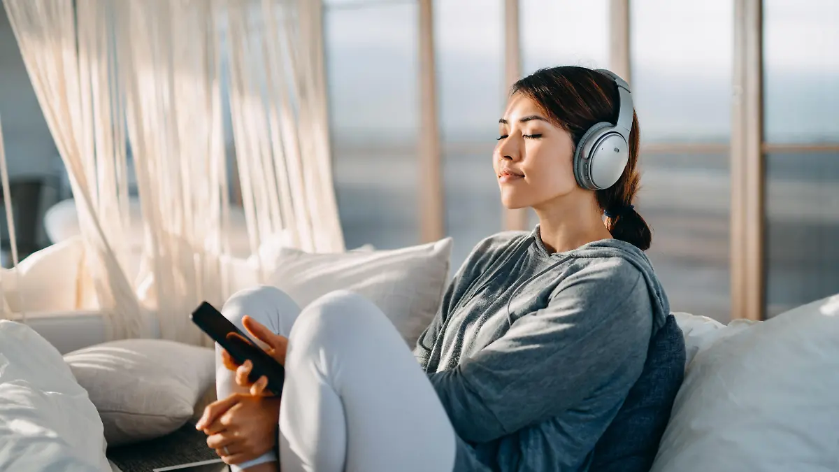 Relaxed young Asian woman with eyes closed sitting on her bed enjoying music over headphones from smartphone at home