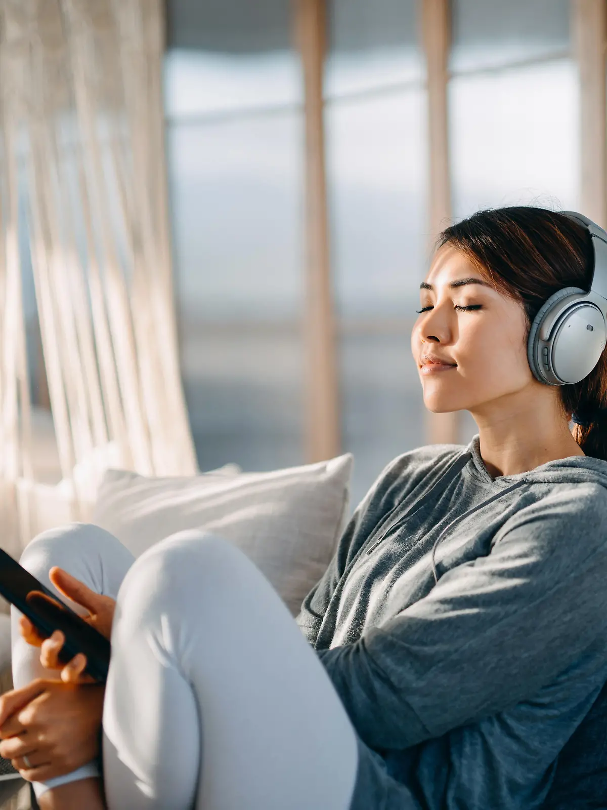 Relaxed young Asian woman with eyes closed sitting on her bed enjoying music over headphones from smartphone at home