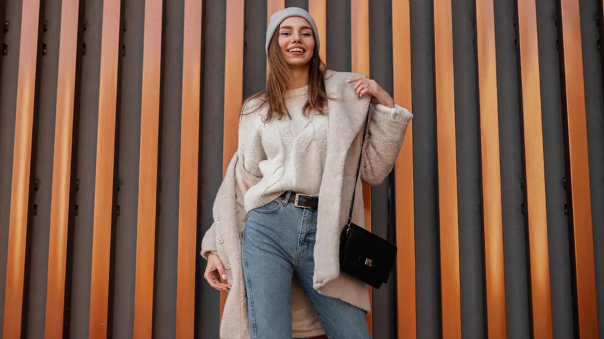 Pretty young happy woman in a warm sweater in a knitted hat in a fashionable faux fur coat in vintage jeans with a leather black handbag stands and smiles near a metal wall. Positive girl model.