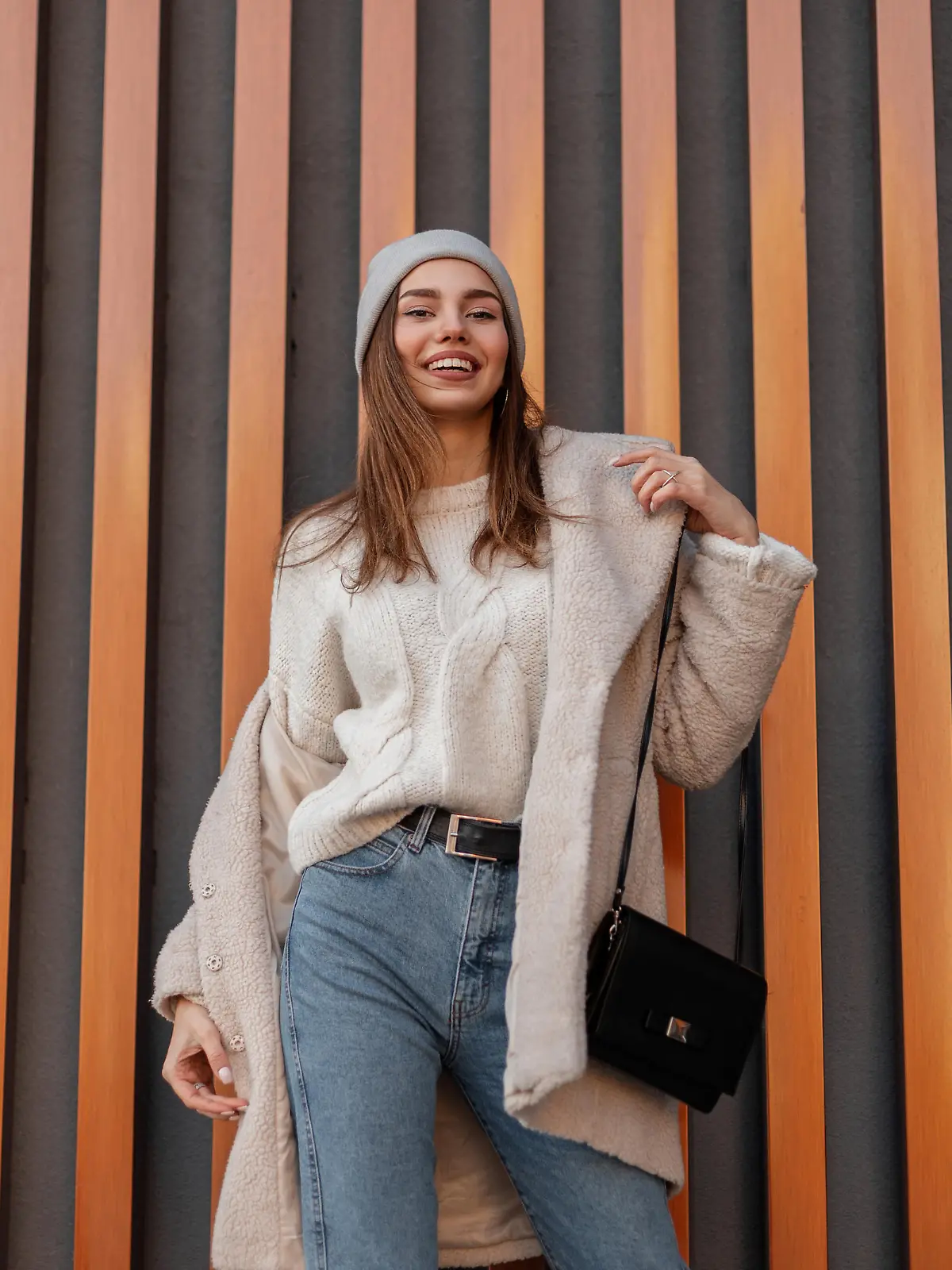 Pretty young happy woman in a warm sweater in a knitted hat in a fashionable faux fur coat in vintage jeans with a leather black handbag stands and smiles near a metal wall. Positive girl model.