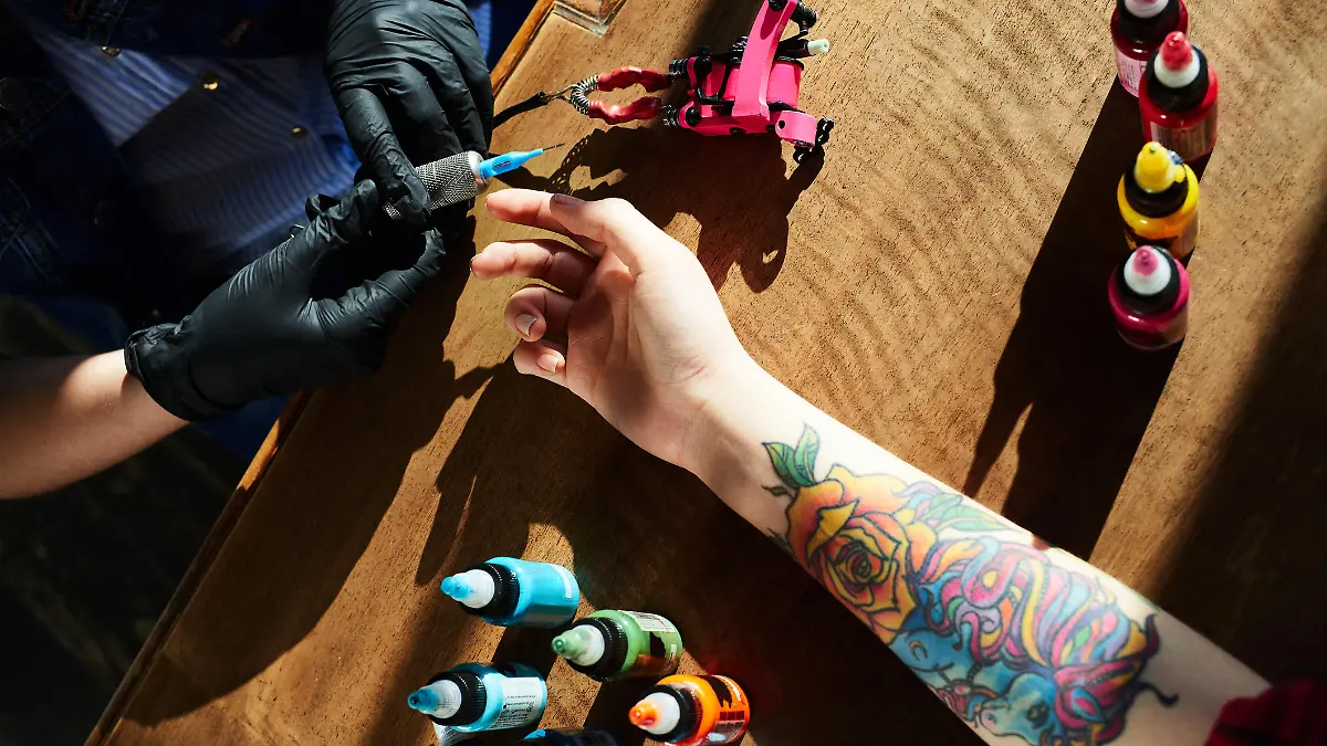 Tattoo expert in gloves holding tattoo gun while hand of female client with new colorful tattoo lying in wooden table beside, high angle close-up view