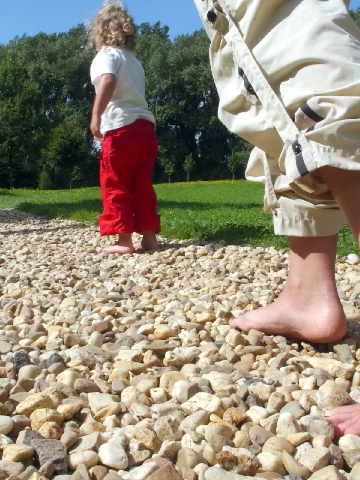 Eine Mutter geht mit ihren beiden Kindern mit nackten Füßen auf groben Kieselsteinen am Freitag (20.08.2004) auf dem ersten Barfuß-Wanderweg am Niederrhein der Stadt Neuß. Auf 300 Metern können sich dort Wanderer barfuß fortbewegen und unterschiedliche Bodenbeläge wie Sand, Steine, Kies, Lehm oder Rindenstücke spüren und so die Natur bewusster erleben. Durch starke und schwache Reize werden die Füsse stimuliert und Muskeln-und Gelenke gestärkt. Foto: Horst Ossinger dpa/lnw