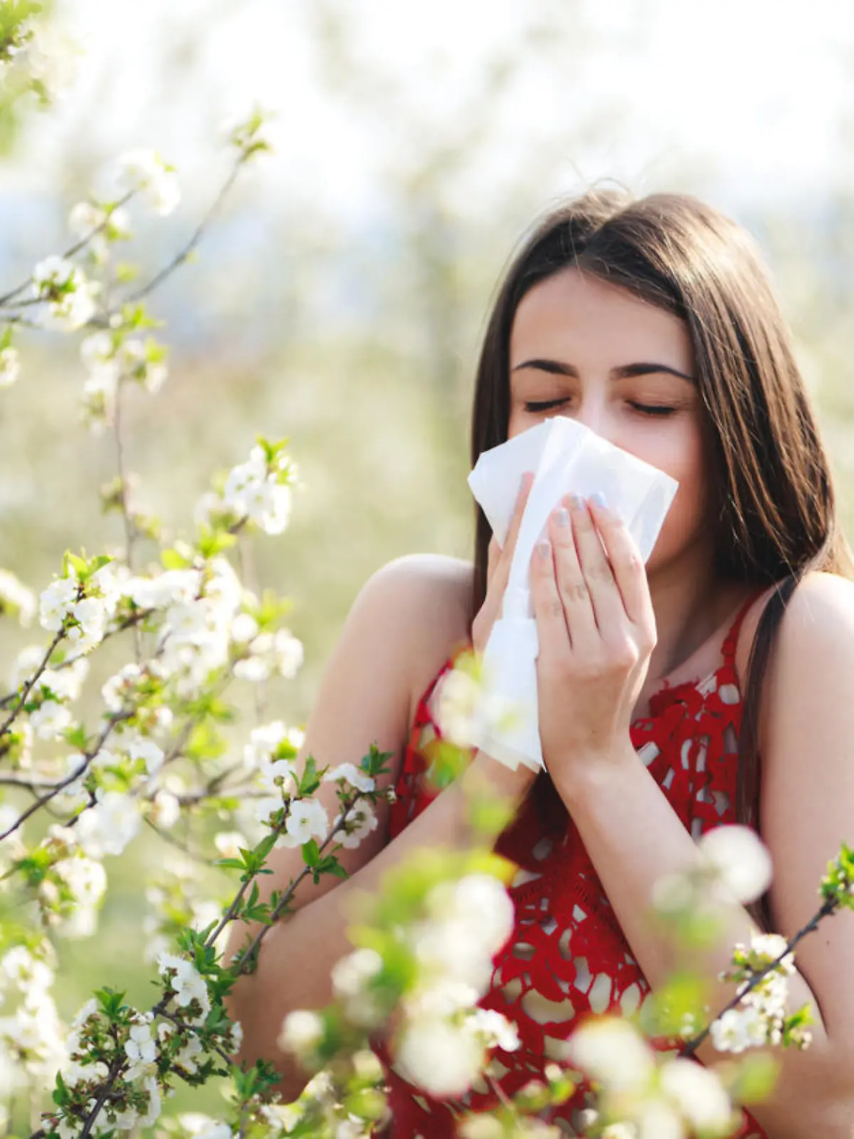 Allergy season, young woman blowing her nose and sneezing in blossoming garden.