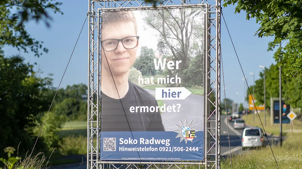 14.06.2021, Bayern, Bayreuth: "Wer hat mich hier ermordet?" steht auf einem großen Plakat mit Foto des Opfers neben dem Tatort. Mithilfe des Plakats sucht die Polizei nach einem Täter, der im Sommer 2020 einen 24-Jährigen in Bayreuth umgebracht haben soll. Die ungewöhnliche Methode sei nach Angaben der Polizei sowohl mit der Familie des Opfers als auch mit der Staatsanwaltschaft abgestimmt. Die Leiche war im August 2020 auf einem Fuß- und Radweg nahe der Universität gefunden worden. Foto: Daniel Karmann/dpa - ACHTUNG: Nur zur redaktionellen Verwendung im Zusammenhang mit dem Fall und nur bis zum Ende der Öffentlichkeitsfahndung der Polizei. +++ dpa-Bildfunk +++