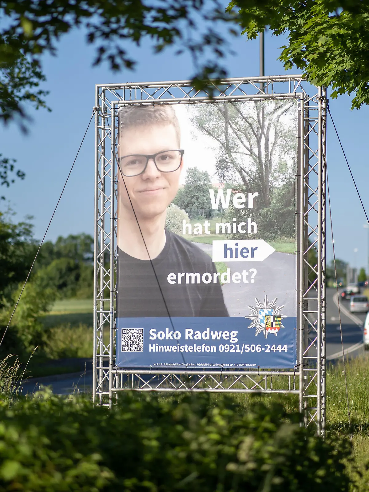 14.06.2021, Bayern, Bayreuth: "Wer hat mich hier ermordet?" steht auf einem großen Plakat mit Foto des Opfers neben dem Tatort. Mithilfe des Plakats sucht die Polizei nach einem Täter, der im Sommer 2020 einen 24-Jährigen in Bayreuth umgebracht haben soll. Die ungewöhnliche Methode sei nach Angaben der Polizei sowohl mit der Familie des Opfers als auch mit der Staatsanwaltschaft abgestimmt. Die Leiche war im August 2020 auf einem Fuß- und Radweg nahe der Universität gefunden worden. Foto: Daniel Karmann/dpa - ACHTUNG: Nur zur redaktionellen Verwendung im Zusammenhang mit dem Fall und nur bis zum Ende der Öffentlichkeitsfahndung der Polizei. +++ dpa-Bildfunk +++