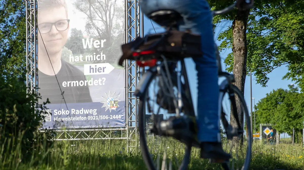 14.06.2021, Bayern, Bayreuth: "Wer hat mich hier ermordet?" steht auf einem großen Plakat mit Foto des Opfers neben dem Tatort. Mithilfe des Plakats sucht die Polizei nach einem Täter, der im Sommer 2020 einen 24-Jährigen in Bayreuth umgebracht haben soll. Die ungewöhnliche Methode sei nach Angaben der Polizei sowohl mit der Familie des Opfers als auch mit der Staatsanwaltschaft abgestimmt. Die Leiche war im August 2020 auf einem Fuß- und Radweg nahe der Universität gefunden worden. Foto: Daniel Karmann/dpa - ACHTUNG: Nur zur redaktionellen Verwendung im Zusammenhang mit dem Fall und nur bis zum Ende der Öffentlichkeitsfahndung der Polizei. +++ dpa-Bildfunk +++