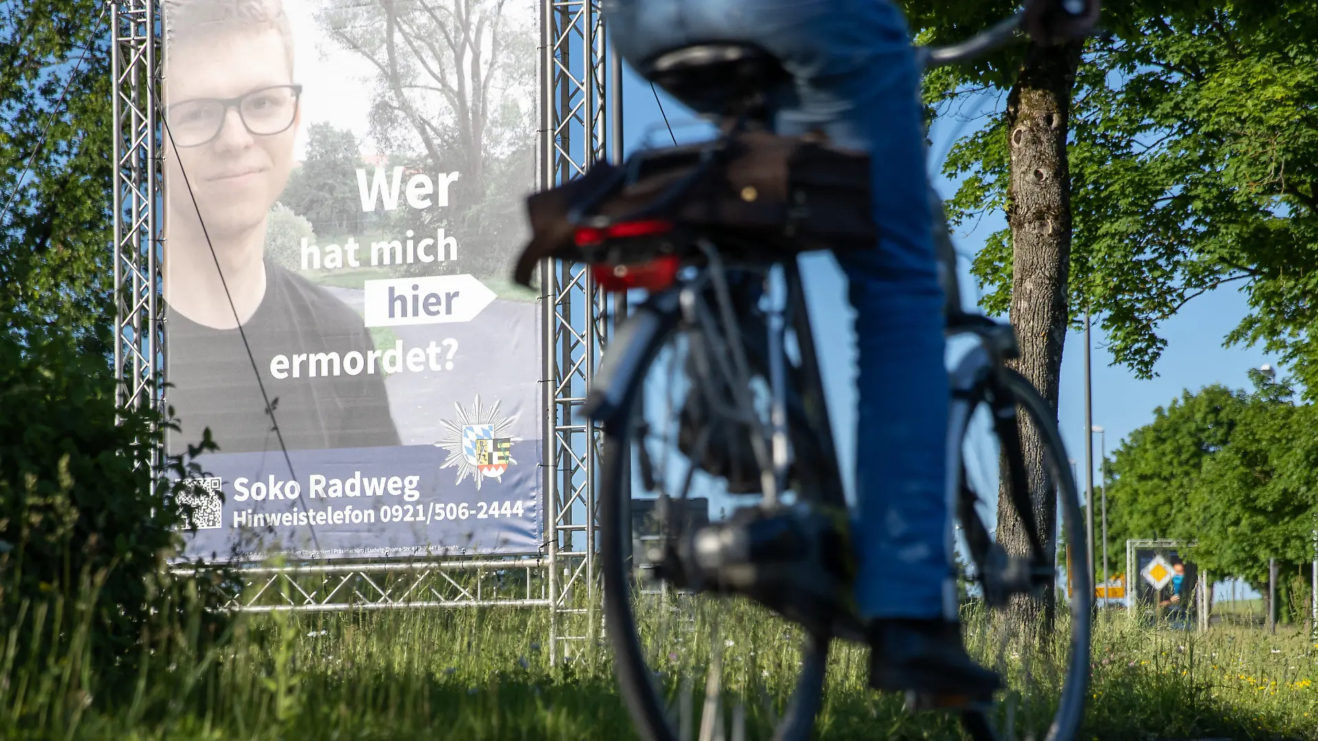 14.06.2021, Bayern, Bayreuth: "Wer hat mich hier ermordet?" steht auf einem großen Plakat mit Foto des Opfers neben dem Tatort. Mithilfe des Plakats sucht die Polizei nach einem Täter, der im Sommer 2020 einen 24-Jährigen in Bayreuth umgebracht haben soll. Die ungewöhnliche Methode sei nach Angaben der Polizei sowohl mit der Familie des Opfers als auch mit der Staatsanwaltschaft abgestimmt. Die Leiche war im August 2020 auf einem Fuß- und Radweg nahe der Universität gefunden worden. Foto: Daniel Karmann/dpa - ACHTUNG: Nur zur redaktionellen Verwendung im Zusammenhang mit dem Fall und nur bis zum Ende der Öffentlichkeitsfahndung der Polizei. +++ dpa-Bildfunk +++