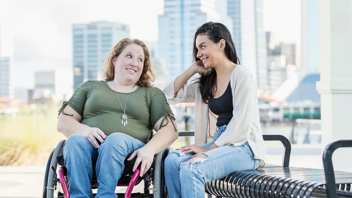Two friends hanging out together on a park bench, city buildings out of focus in the background. One of them, a young woman in her 20s, is sitting in a wheelchair. She was born with spina bifida. A mid adult Hispanic woman is sitting on the bench beside her. They are smiling and looking at each other.