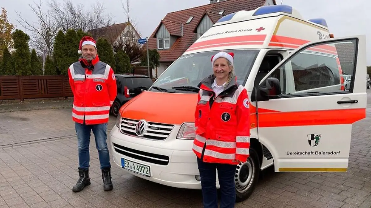 Zwei Mitarbeiter des Roten Kreuzes stehen mit Weihnachtsmütze vor einem Rettungswagen.