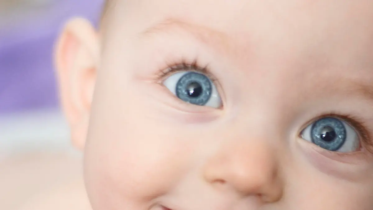 A close up of a baby girl's face as she smiles at the camera. She is 10 months old. She has two bottom front teeth and big blue eyes. A light purple towel behind her is out of focus due to the shallow depth of field. She has bare skin because it is bath time.