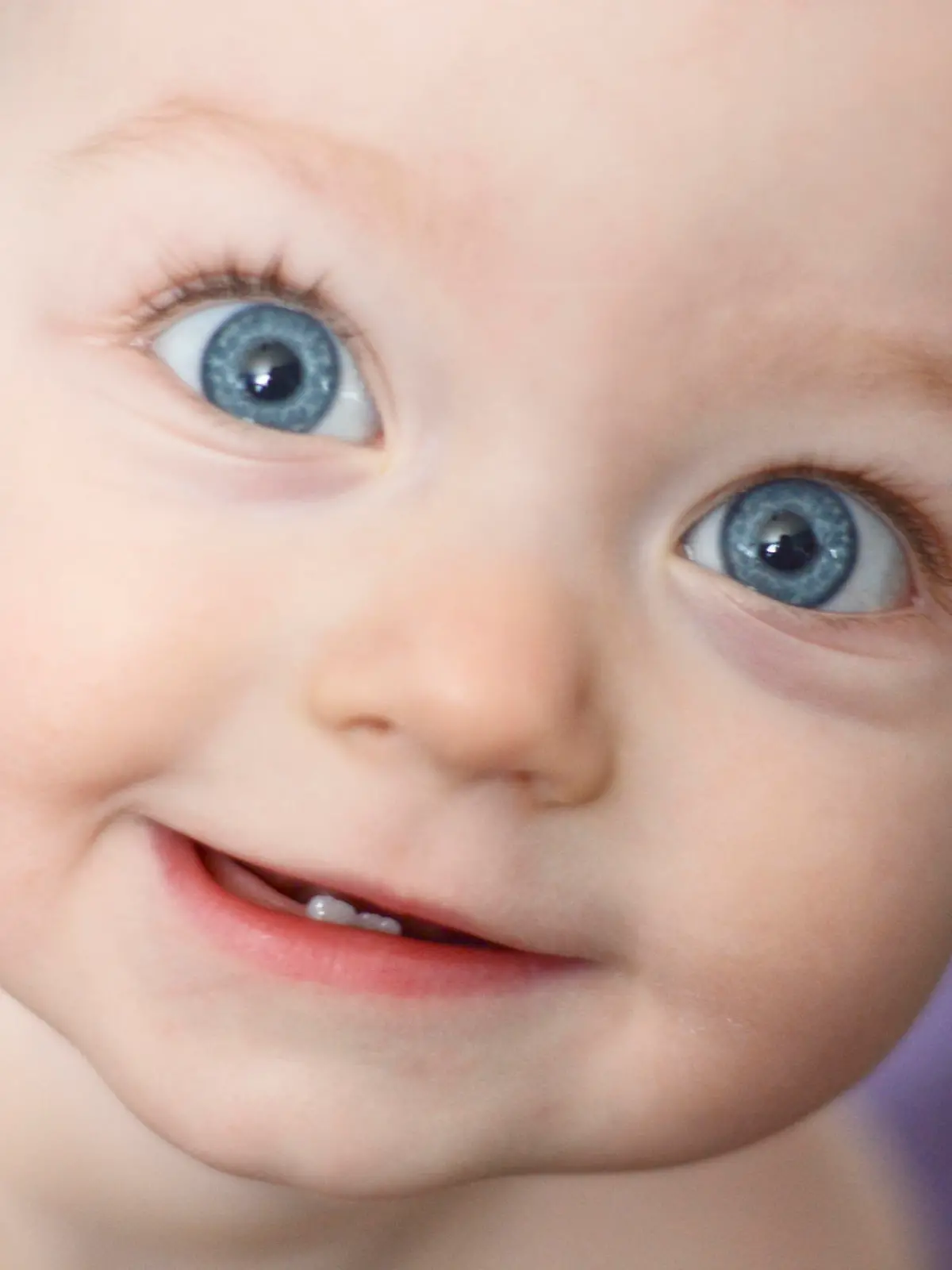 A close up of a baby girl's face as she smiles at the camera. She is 10 months old. She has two bottom front teeth and big blue eyes. A light purple towel behind her is out of focus due to the shallow depth of field. She has bare skin because it is bath time.