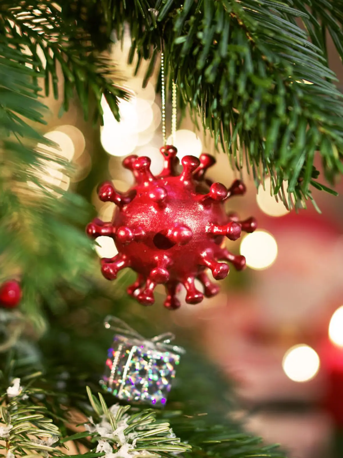 A Christmas Ball hanging on a decorated christmastree with candels in the background. The Christmas Ball is shaped like a Corona virus.