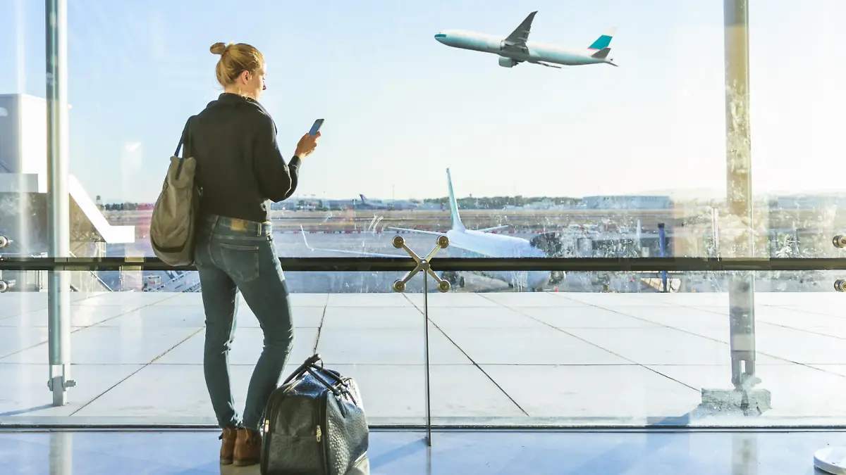 Young casual female traveler at airport, holding smart phone device, looking through the airport gate windows at planes on airport runway. || Modellfreigabe vorhanden