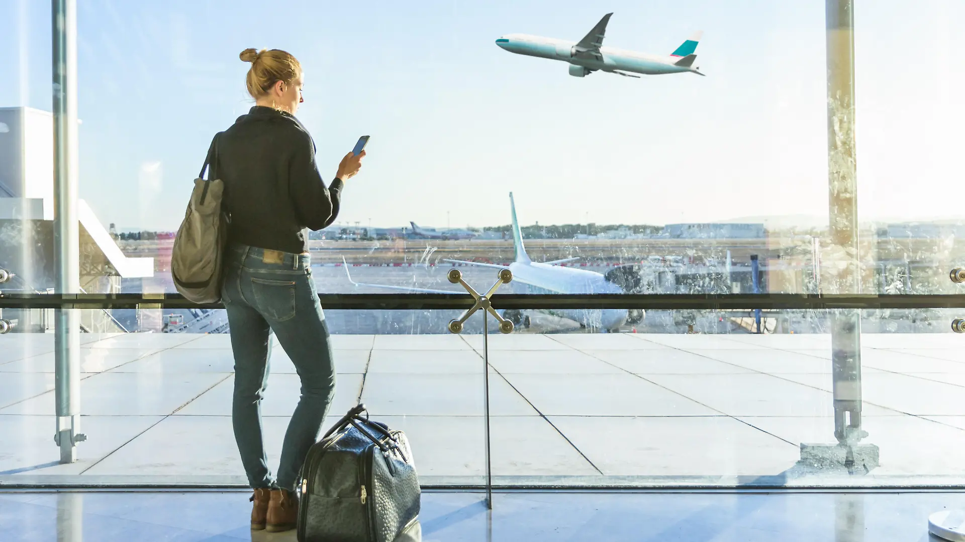 Young casual female traveler at airport, holding smart phone device, looking through the airport gate windows at planes on airport runway. || Modellfreigabe vorhanden