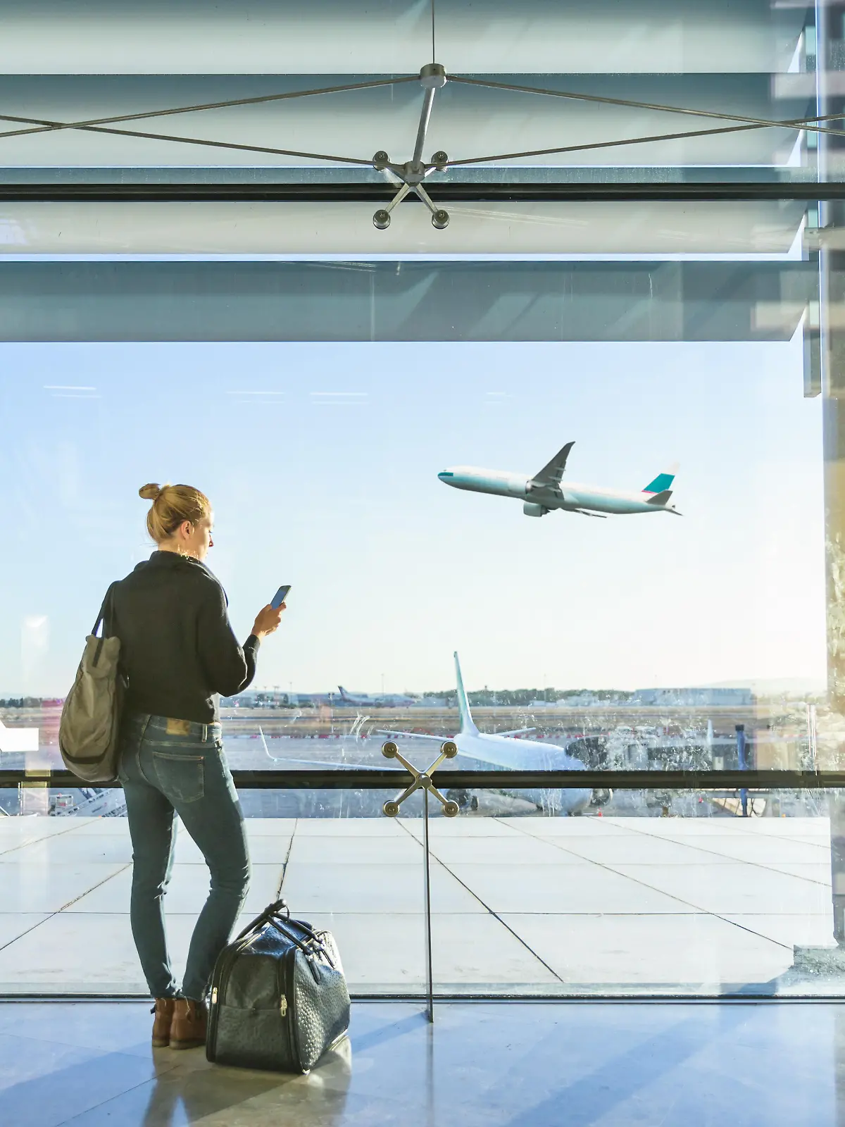 Young casual female traveler at airport, holding smart phone device, looking through the airport gate windows at planes on airport runway. || Modellfreigabe vorhanden